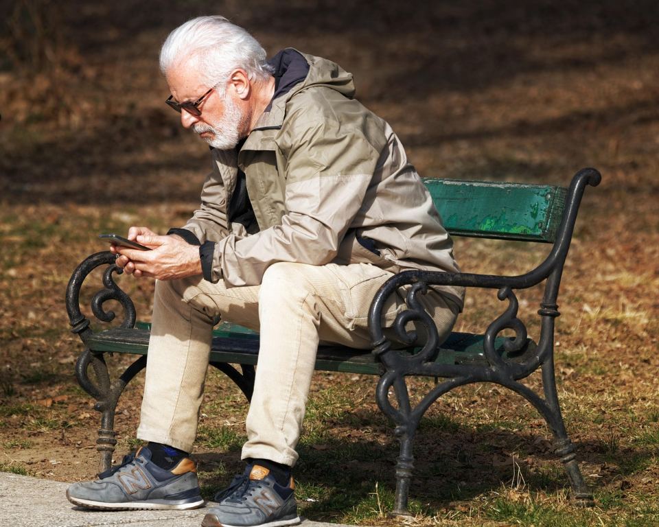 Senhor de cabelo branco sentado num banco de praça, mexendo no celular
